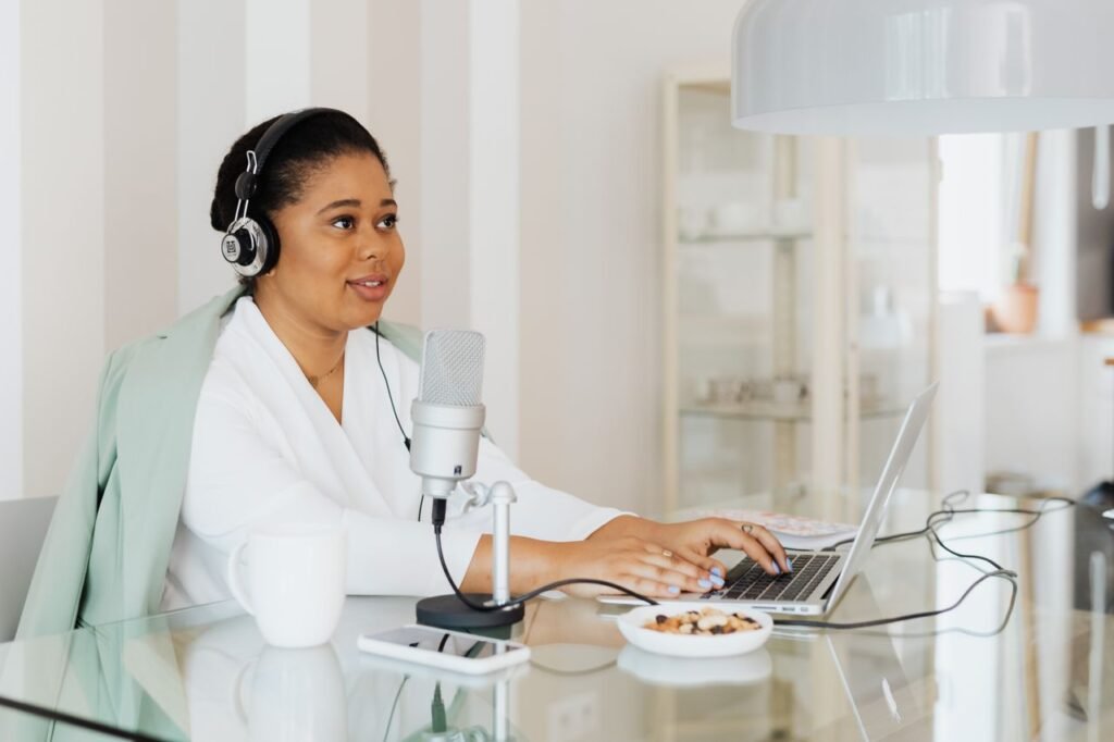 A woman working on a podcast in a modern home studio setup, using a laptop and microphone.